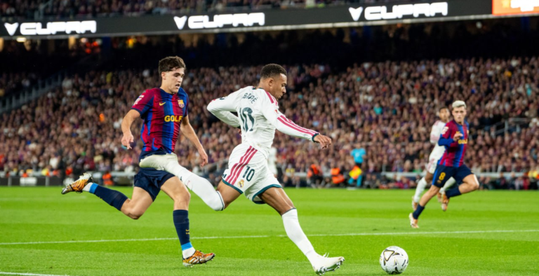 Kylian Mbappé Real Madridin valkoisessa peliasussa kurottaa jalkaansa palloa kohti Barcelonan pelaajan ollessa vieressä.
